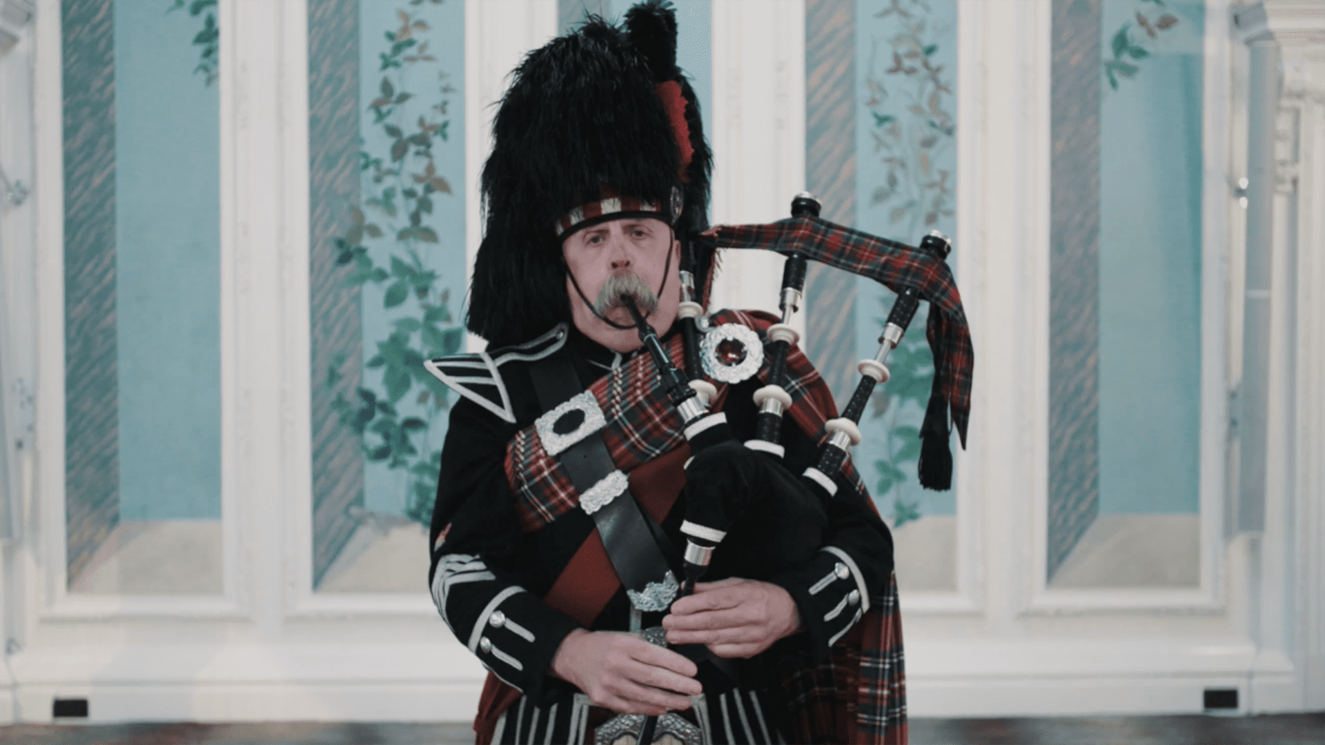 Man with moustache playing bagpipes while dressed in Scottish uniform